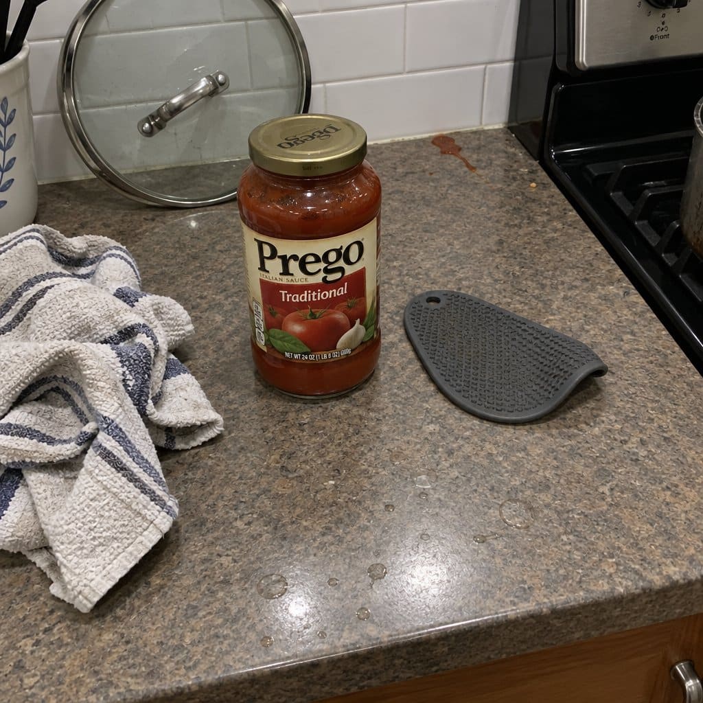 An unopened pasta sauce jar sitting on a kitchen counter next to a worn rubber jar-grip pad, a dish towel and pot lid nearby