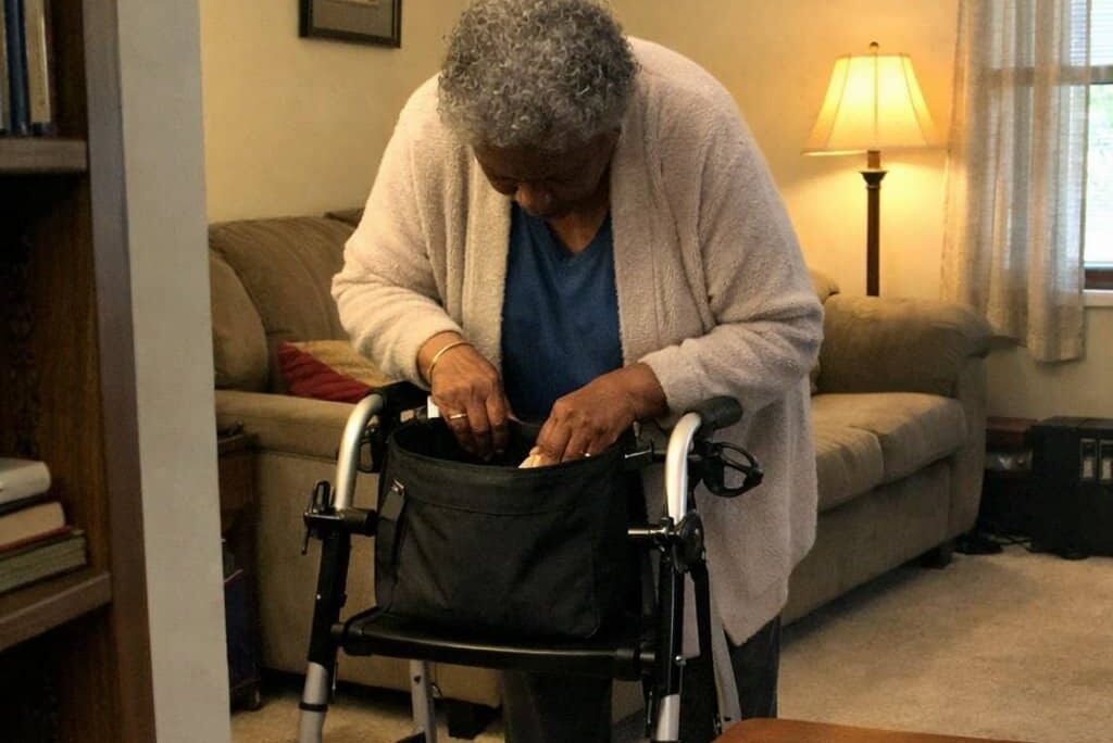 An older woman standing at her walker reaches into a bag mounted across the front crossbar, inside a lived-in living room with a lamp and side table nearby