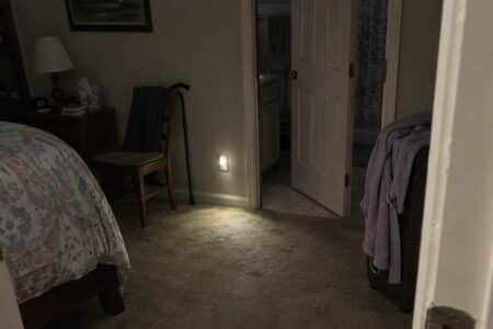 An empty dim hallway between bedroom and bathroom with a low light on the wall and slippers and a cane nearby.