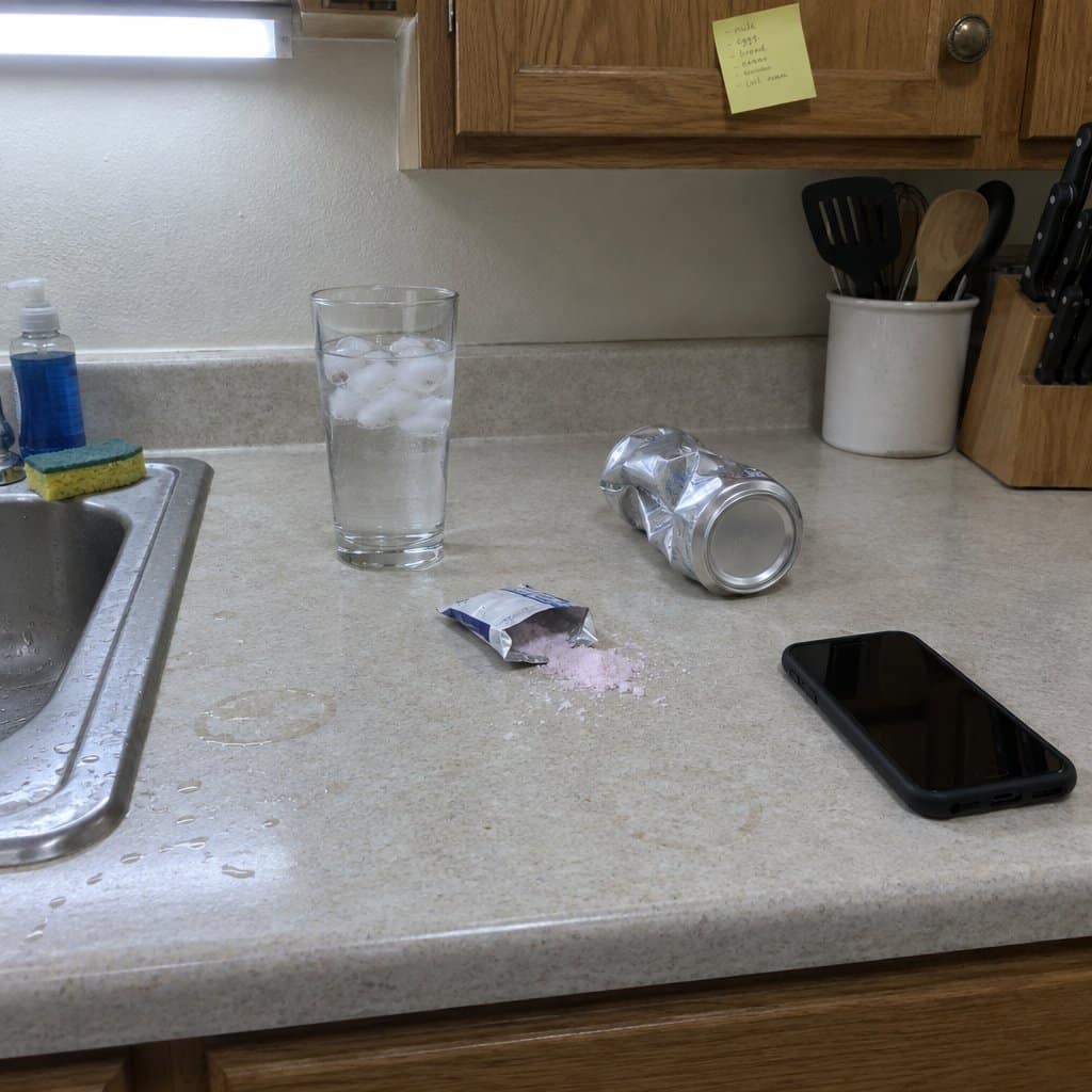 A kitchen countertop with a tall glass of water with ice, an open powder packet, a crumpled energy drink can turned label-side down, and a dark cell phone screen, overhead light on