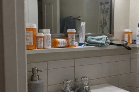 A narrow bathroom shelf crowded with prescription bottles of different sizes, one tipped on its side, with a folded washcloth and other items