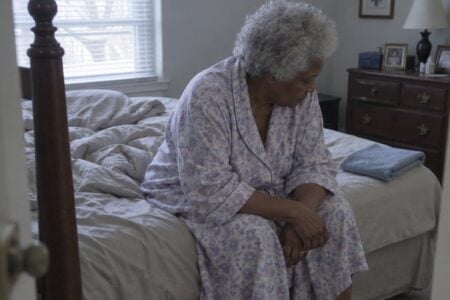 An older woman with natural grey-white hair sits on the edge of a bed with elbows on knees, looking downward, slippers on the floor and a folded towel on the mattress beside her