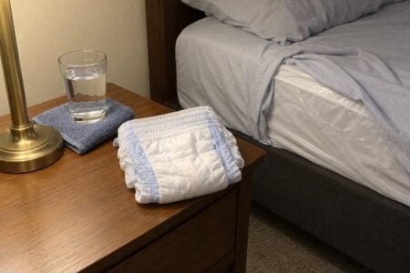 Overnight pull-on briefs folded on a nightstand beside a glass of water, with a mattress protector visible at the edge of the bed