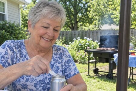 Older woman at a picnic table opening a soda can with a small plastic lever tool on a sunny patio